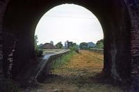A westward view through one of the three road bridge arches at Bungay station in June 1976. This station had been closed to all traffic in 1964 and the remains seen here, including the bridge, were swept away in the early 1980s when the A143 was rerouted along the trackbed. The brick column visible is the former water tower base, the tank which used to rest on it having been salvaged for use at Weybourne on the North Norfolk railway.<br><br>[Mark Dufton 19/06/1976]