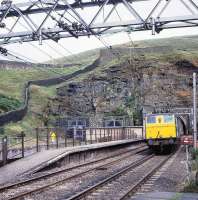 EM1 no 76046 exits the western portal of Woodhead Tunnel with a freight on 15 August 1979. The train is about to pass the marker board  for platform 4 at Woodhead station, which had closed to passengers in July 1964. Built in 1952 as 26046, the locomotive carried the name <I>Archimedes</I> until the late 60s and was withdrawn in November 1980. <br><br>[Peter Todd 15/08/1979]