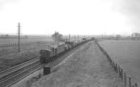 An EE Type 4 takes a freight north from Newcastle in the 1960s, near the site of Little Benton sidings. The train has recently crossed the bow-string girder bridge over the Coast Road in the background. Also in view is the eastern end of the WD&HO Wills cigarette factory, the main module of which still stands, having been converted into luxury flats after closure. Also of note alongside the down line in the foreground is a fogman's lever used for the remote placement of detonators, suggesting the train is approaching a distant signal [see image 30435].<br><br>[K A Gray //]