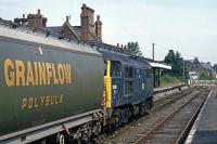 A train of empty polybulks squeaks and clanks to a halt at Dereham in July 1987. Running several hours late, it is heading for the granary at North Elmham. One of the crew of 31 106 has now to brave the stifling midday heat and swarms of thrips to open the crossing gates.<br><br>[Mark Dufton /07/1987]