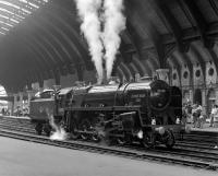 The curved trainshed at York forms a magnificent backdrop to 9F No. 92220 <I>Evening Star</I> blowing-off impatiently as it waits to take over the Scarborough Flyer excursion in July 1976. The train started its journey from Newcastle round the Durham coast behind Stephenson valve gear Black 5 No. 4767, with class 40 diesel power taking over at Stockton for the second leg along the ECML.<br><br>[Bill Jamieson 04/07/1976]