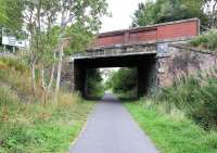 Looking east along the Peebles loop near the site of Rosewell & Hawthornden station (just behind camera) on 4 September 2013. The platforms survive here, though are now totally overgrown [see image 13813]. This bridge over the trackbed links the east end of Rosewell with the A6094 that now bypasses the village. As indicated by the road sign, the view is towards Bonnyrigg.<br><br>[John Furnevel 04/09/2013]