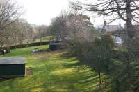 A view east from the road bridge that once crossed the railway at Westgate-in-Weardale, photographed on 19 March 2015. The station house is on the right but less clear is the former station building, painted grey, hiding in the trees in the middle of the picture beyond the wall running left to right across the trackbed. [Ref query 7589]<br><br>[John McIntyre 19/03/2015]