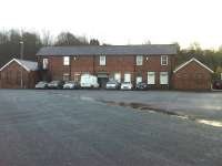 A view from the adjacent road showing the large but rather bland frontage to Pontefract Baghill Station. The left half of the main building and the single storey wing are in use as a driving test centre, whilst the right side is in occasional railway use. The entrance to the platforms is through a small tunnel in the centre of the main building, with a replica of the station running in board mounted over the tunnel entrance.<br><br>[David Pesterfield 13/11/2015]