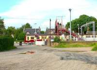 Passengers exit Cardross station via the pedestrian footbridge on 27 July 2005, having recently disembarked from a Helensburgh bound train. The motor cyclist is forced to wait for the train to clear the level crossing before recommencing his journey. View north along Station Road towards the junction with the A814. <br><br>[John Furnevel 27/07/2005]