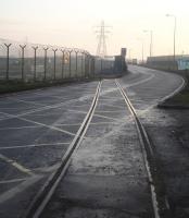 The disused and severed spur from the Sheerness branch that formerly served the Sheerness Steel site (beyond the fence to left) and continued on to Sheerness Docks. View is south along the A429 Brielle Way in January 2017. The line to the docks is blocked some 100m behind the camera and the section of trackbed running beyond the crossing to the former South Junction is currently being excavated. There are plans to re-open part of the steelworks later in the year, initially as a rolling mill. It is possible the connection between the branch and the steelworks may yet be restored. [Ref query 654]<br><br>[David Pesterfield 24/01/2017]