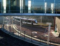 A tram swings left after running below the A8 on 26 January 2017 on the approach to Edinburgh Gateway station with an airport bound service. View is from the upper level of the station, showing part of the elevated covered walkway which links the main concourse and the tram platforms via escalators situated off to the right. The lines in the foreground continue behind the station and into Gogar tram depot.<br><br>[John Furnevel 26/01/2017]