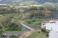 A passenger train on the St Kitts Scenic Railway, running north along the east coast, swings inland round the head of one of the numerous valleys. The first vehicle behind the 0-6-0DH is a generator car that powers services on the five coaches. The truck about to pass under the viaduct is a support vehicle that shadows the train with the driver operating the level crossing barriers. 180217.