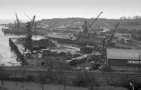 View from a passing train of the R & M Supplies (formerly T W Ward) scrapyard at Inverkeithing Bay.  In the foreground is the Rosyth Dockyard branch.  A Fowler diesel shunts cement wagons with many more in the background.  These were of a batch involved in a derailment at Cupar on 4 July 1988 which identified an axle flaw as the cause.  The Whitby class frigate HMS Eastbourne F73 nears the end by the jetty.  1989.<br><br>[Bill Roberton //1989]